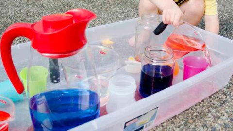 A child in a yellow shirt leans over a storage container full of cups and water. He's pouring orange water into a cup, and missing.