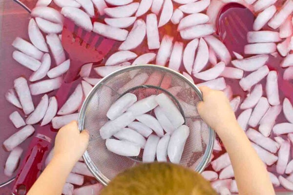 Child playing in a storage container (sensory bin) with ice cubes floating in red-dyed water. Kitchen tools and colanders are in the image.