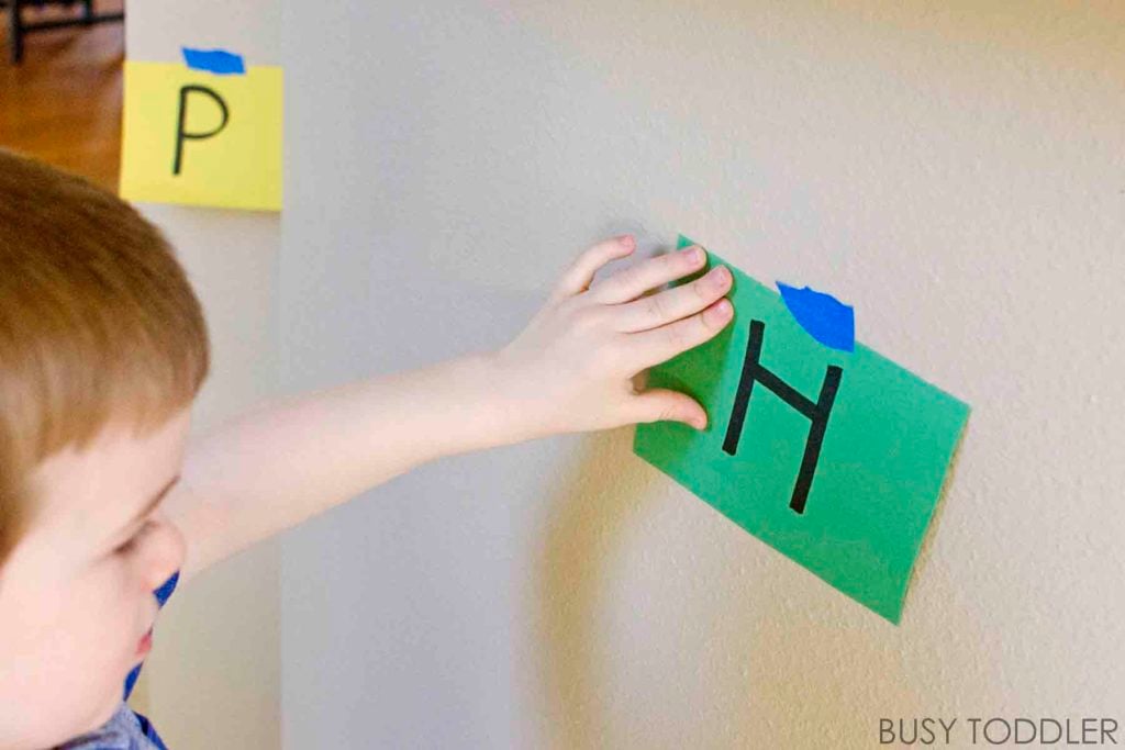 A preschooler touches the letter H on green construction paper that has been taped to a wall.