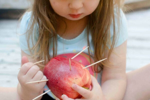 A toddler holds a red apple and a toothpick. She is sticking the toothpicks into the apple.