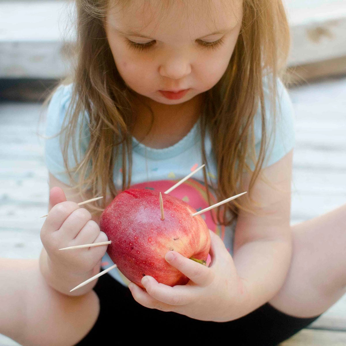 A toddler holds a red apple and a toothpick. She is sticking the toothpicks into the apple.