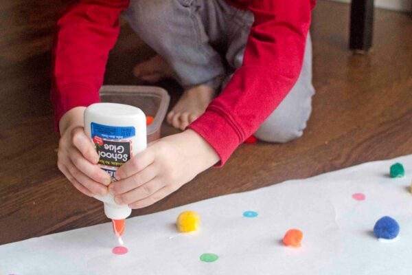 A child crouched over a long strip of butcher paper covered in colorful dots, gluing a pom pom onto a matching dot.