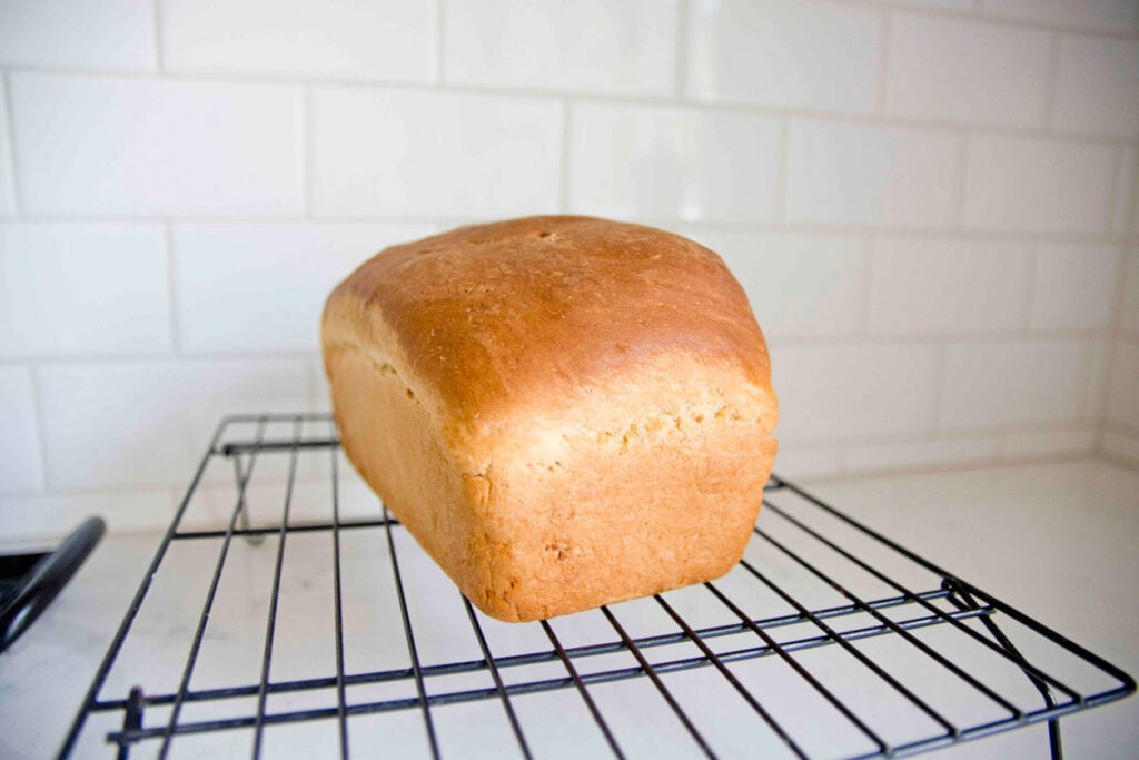 A loaf of bread sits on a wire cooling rack with subway tiles in the background.