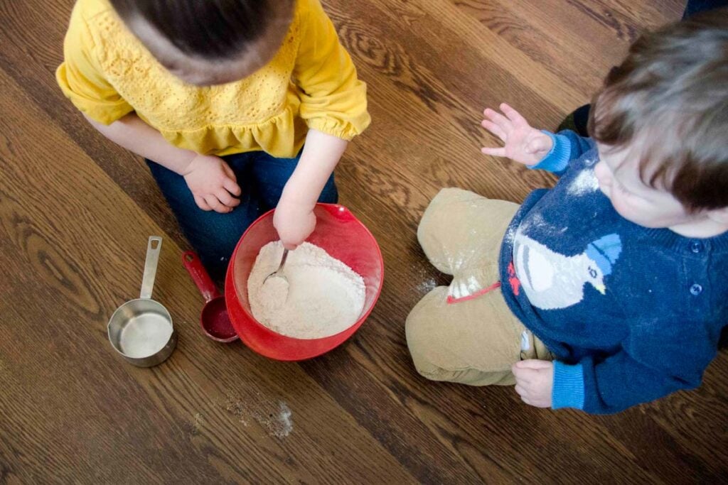 A 1.5 year old and 3 year old mix bread ingredients in a bowl.