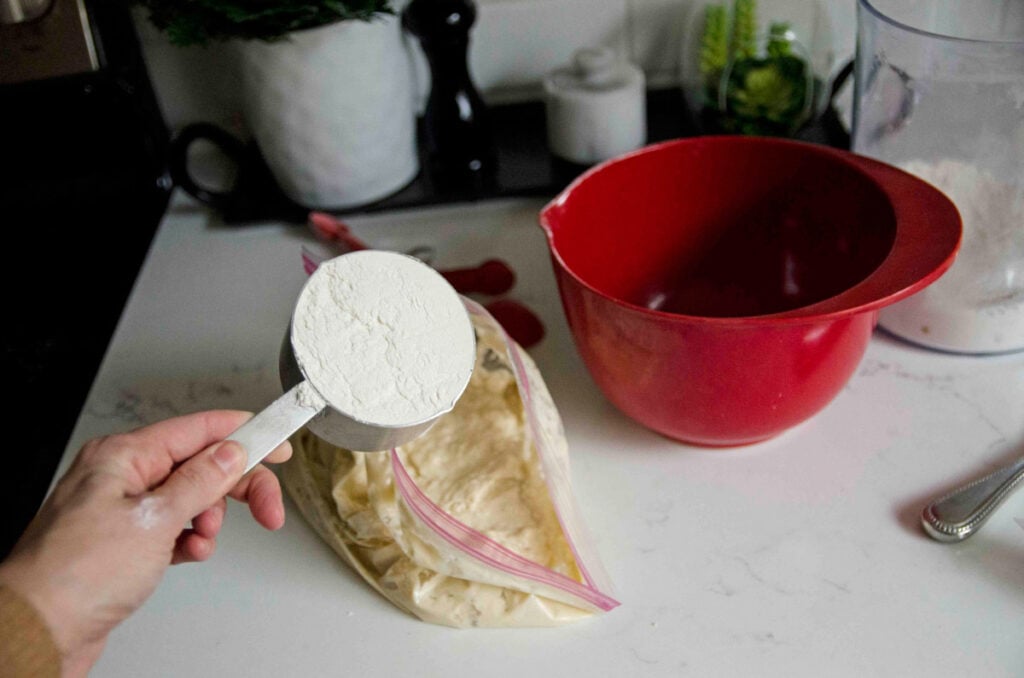 A woman's hand adds a cup of flour to a bag of bread dough.