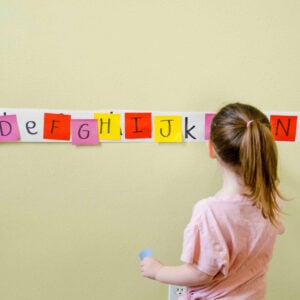 A toddler holds a blue sticky note looking at a long strip of lower case letters. She is matching upper case letters to them.