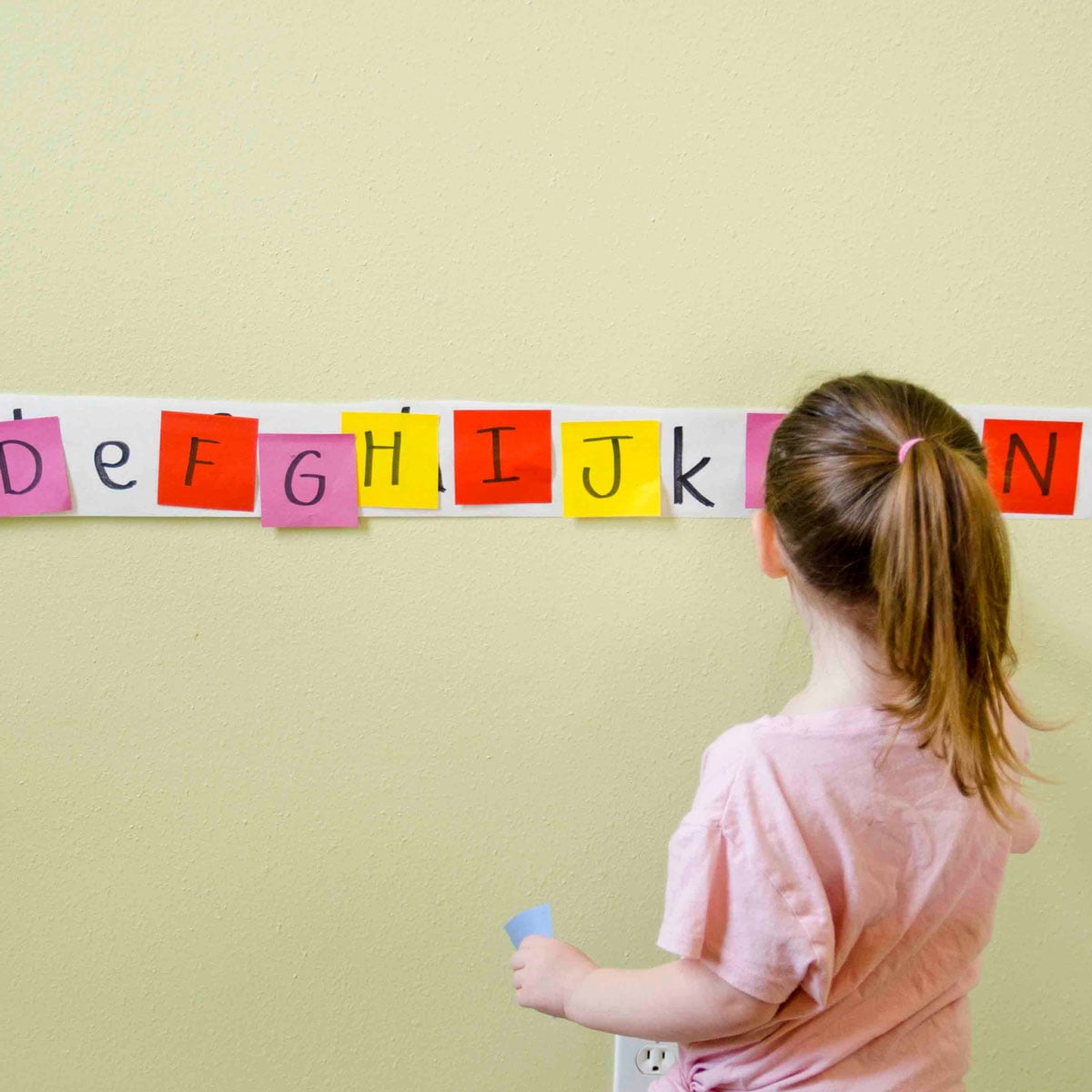 A toddler holds a blue sticky note looking at a long strip of lower case letters. She is matching upper case letters to them.
