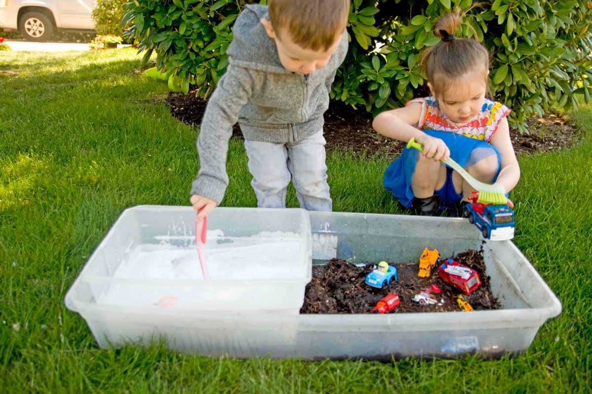 A toddler and preschoolers sit on grass with a sensory bin full of mud in front of them. They are cleaning toy cars with soapy water and scrub brushes.
