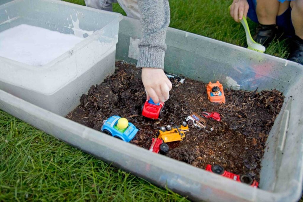 A toddler's hand pushes cars in a muddy car sensory bin.