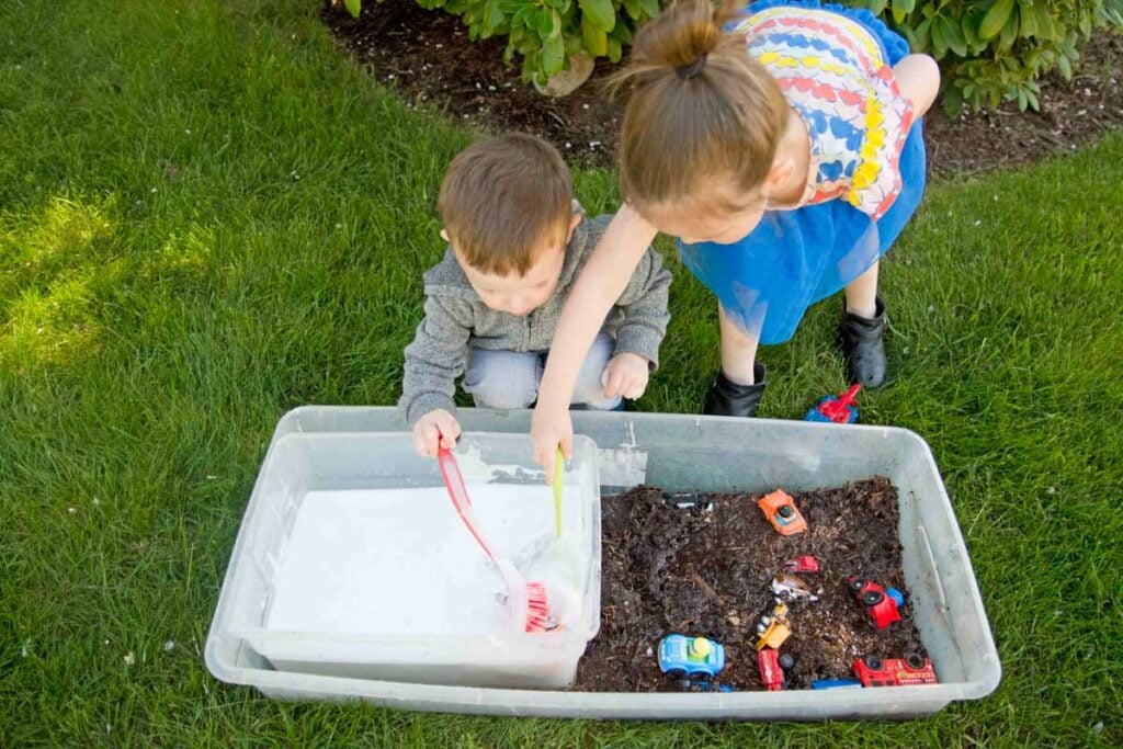 A toddler and preschoolers sit on grass with a sensory bin full of mud in front of them. They are cleaning toy cars with soapy water and scrub brushes.