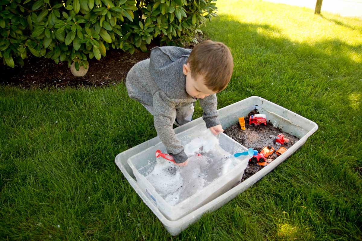 A toddler sloshes muddy water as he cleans cars in an outdoor sensory bin.
