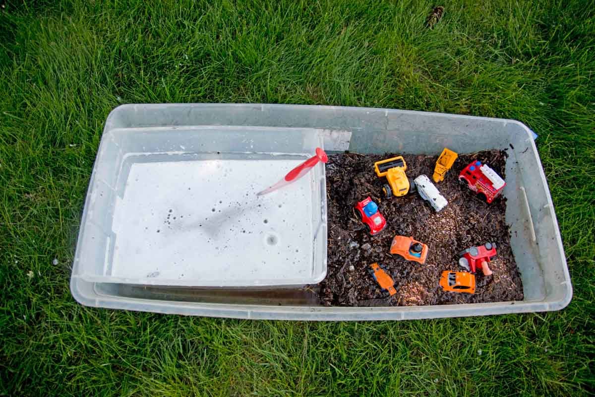 A sensory bin of mud and toy trucks sits on grass. A second sensory bin with soapy water sits with it.