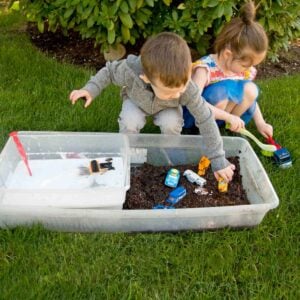 A toddler and preschoolers sit on grass with a sensory bin full of mud in front of them. They are cleaning toy cars with soapy water and scrub brushes.