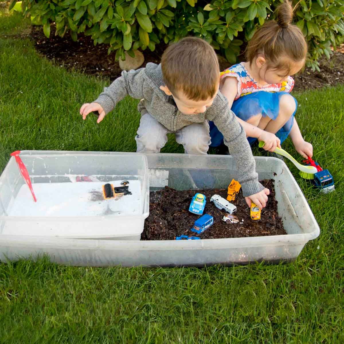 A toddler and preschoolers sit on grass with a sensory bin full of mud in front of them. They are cleaning toy cars with soapy water and scrub brushes.