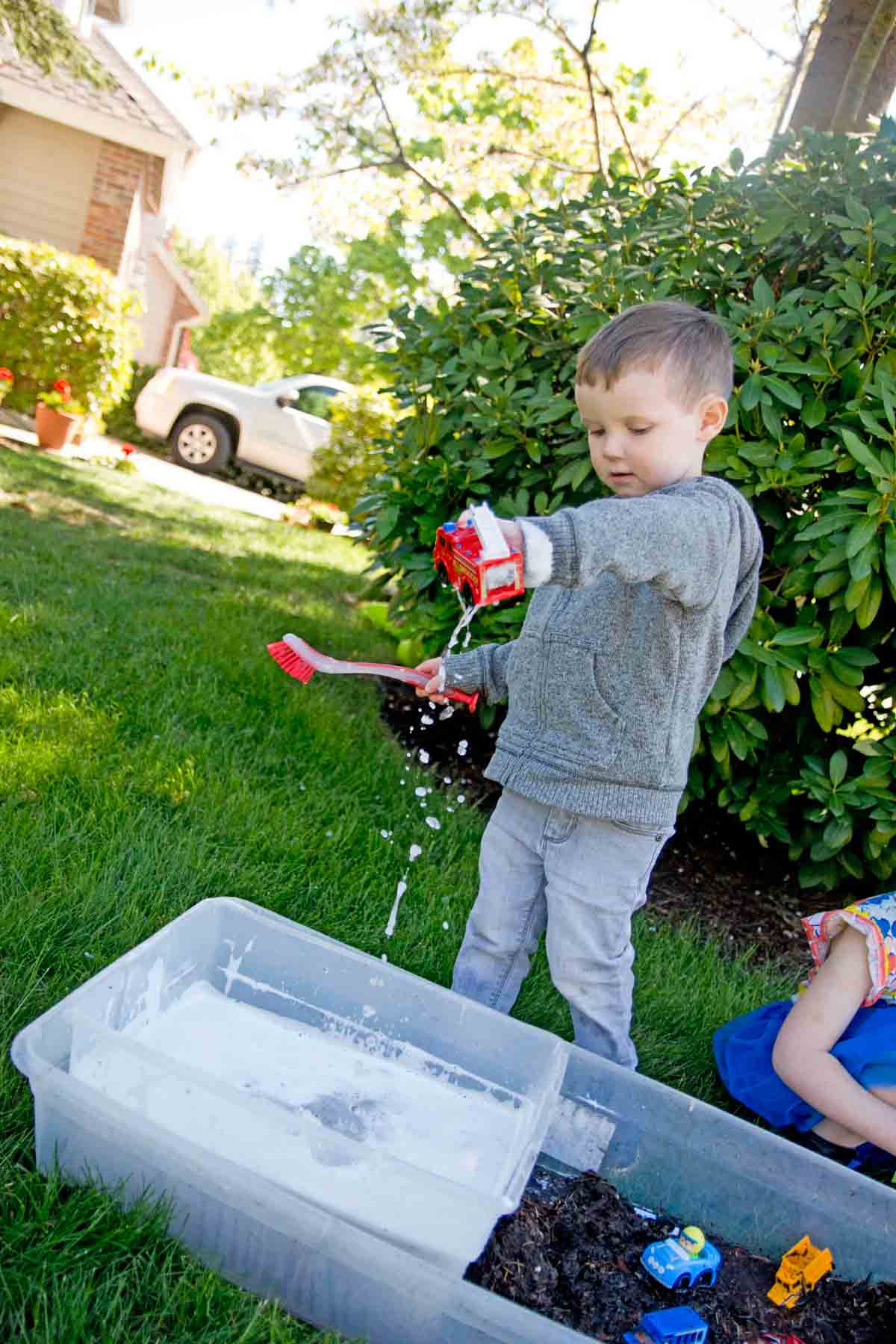 A toddler stands and lets water drip from a plastic fire truck while playing outside in a muddy sensory bin.