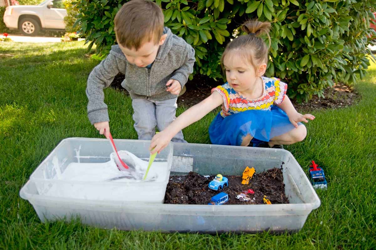 A toddler and preschoolers sit on grass with a sensory bin full of mud in front of them. They are cleaning toy cars with soapy water and scrub brushes.