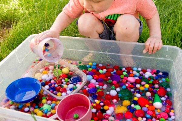 A child pours pom pom balls back into a sensory bin that is full of water.