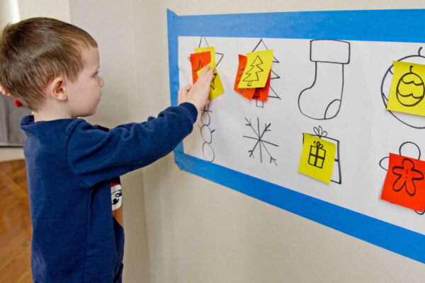 A child in a blue sweater places a sticky note onto a large sheet of white paper. Drawn on the paper is Christmas images.