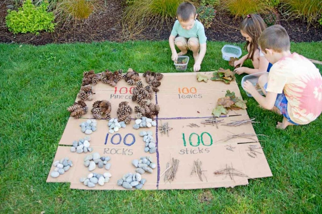Three kids sit around a flat box making groups of 10s to 100 of nature items.