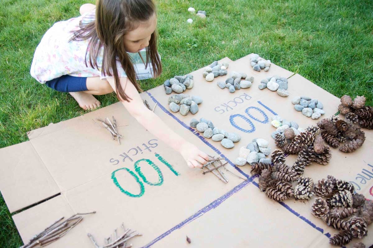 A child learns over a flat box, making piles of 10 sticks to count to 100. 100 rocks and pinecones are also visible.