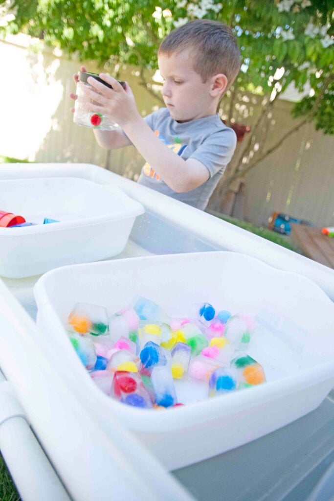 A child mixes frozen pom pom balls into a plastic container with water and shakes it.