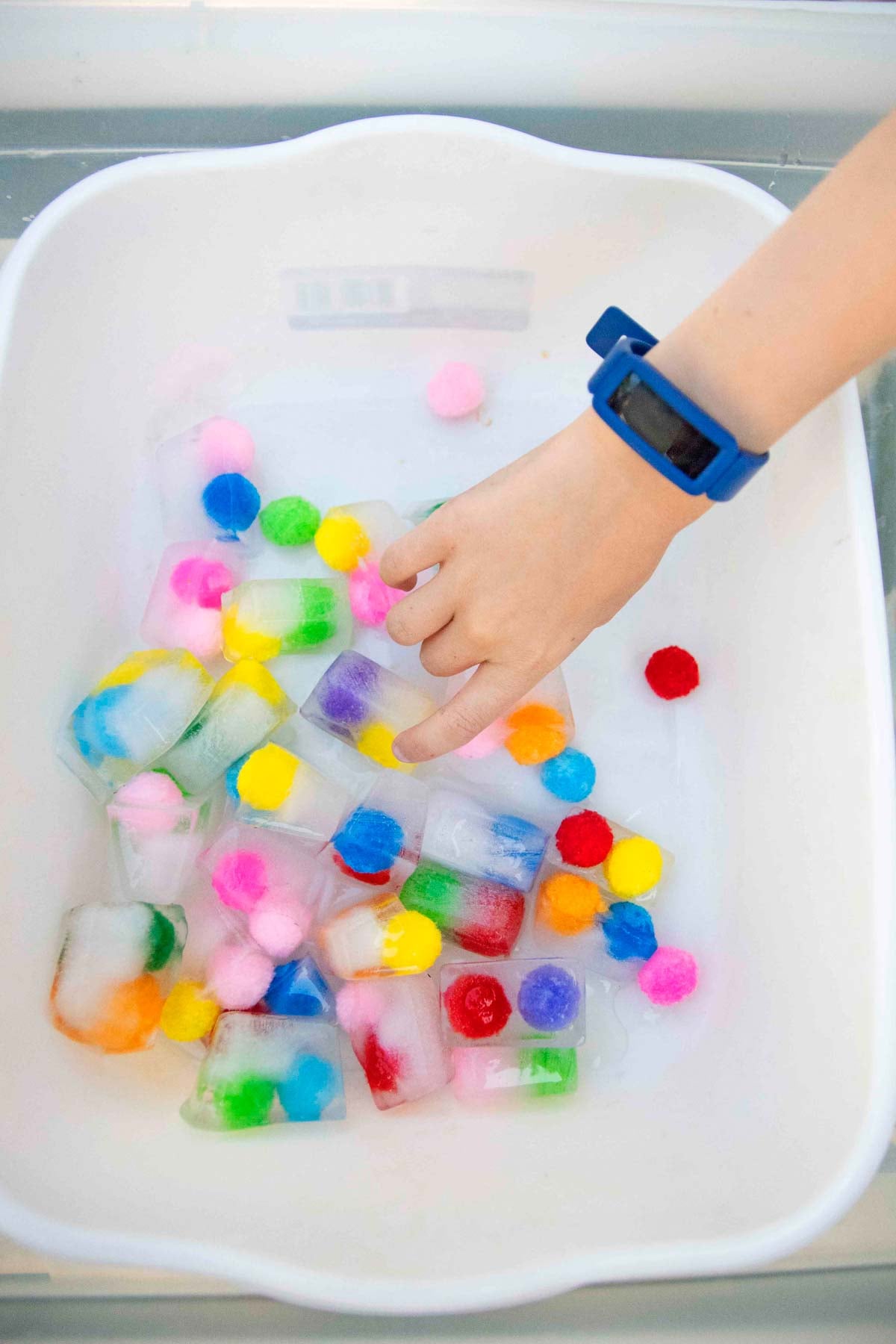 A child's hand grabs a frozen pom pom ball ice cube from a white tub.