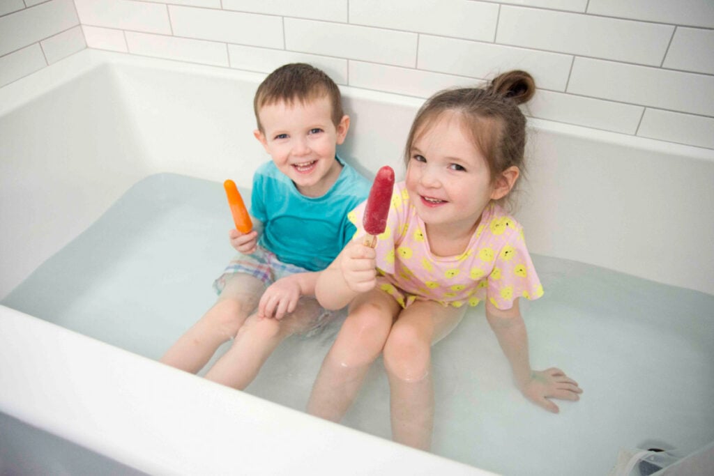 Two kids sit in a bathtub with swim suits on. They are eating popsicles.
