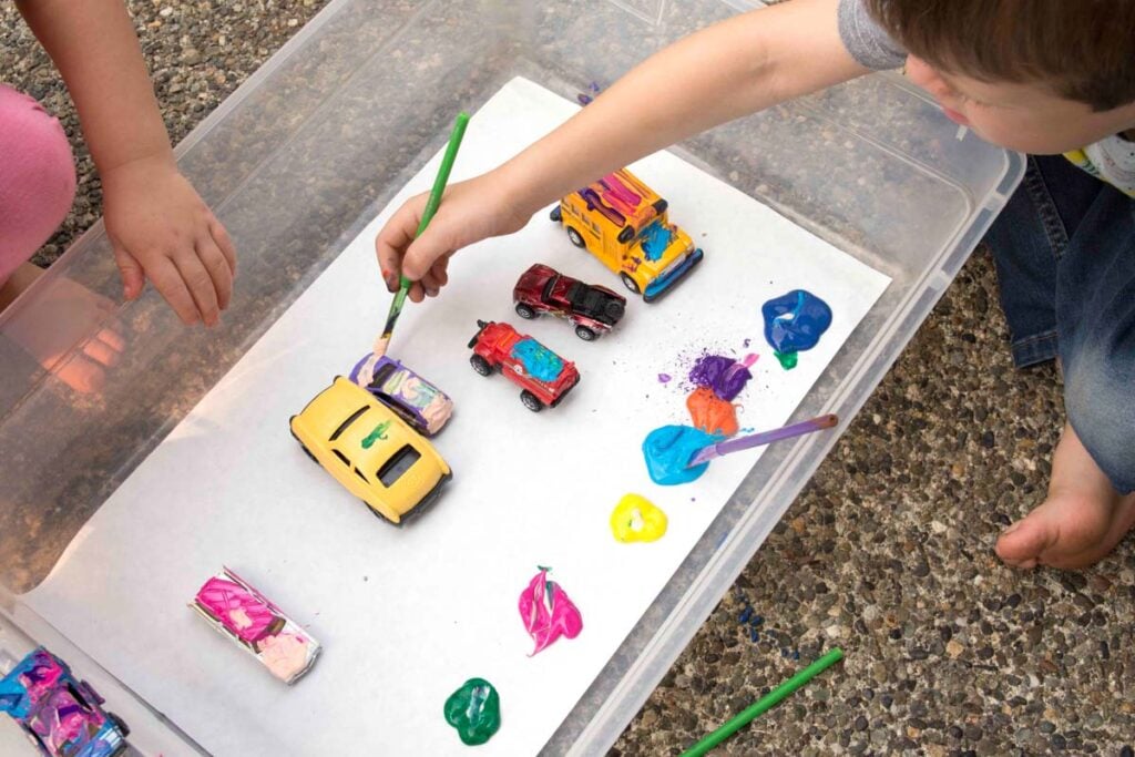 Two children sit outside holding paint brushes and metal cars. Many cars are sitting in a storage container. The children are painting the cars.