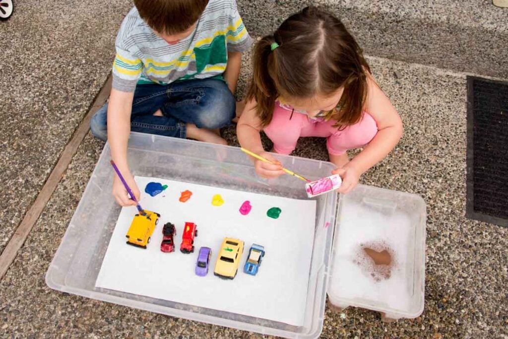 Two children sit outside holding paint brushes and metal cars. Many cars are sitting in a storage container. The children are painting the cars.