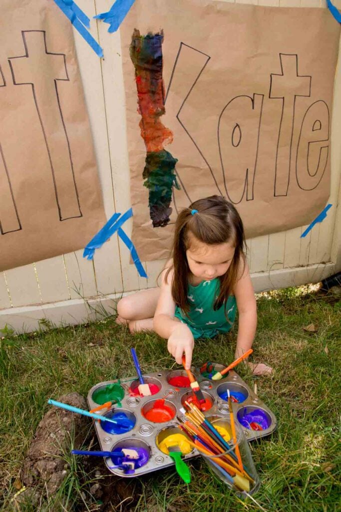 A child dips a brush into paint in front of a fence. On the fence is paper with their name (KATE) printed. 