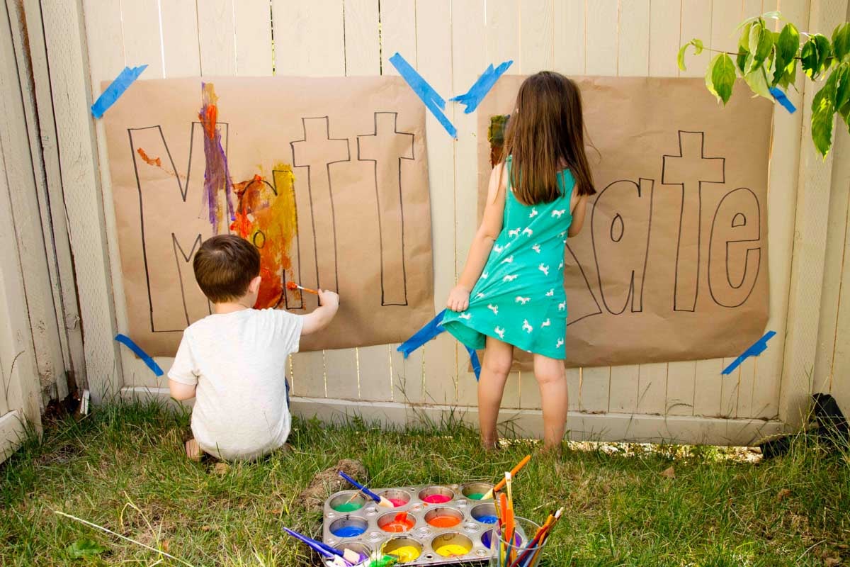 Two kids (2 and 4) standing at large pieces of paper with their names drawn on them beginning to paint.