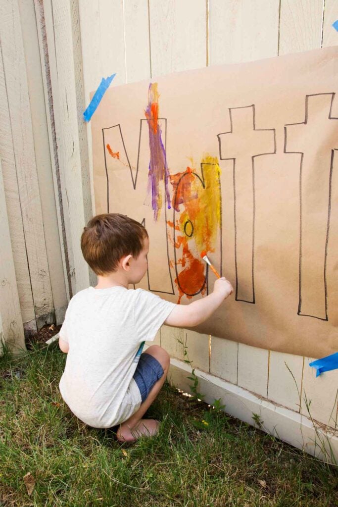 A child squats on grass in front of a fence. On the fence is paper with their name (MATT) printed. The child is painting the paper.