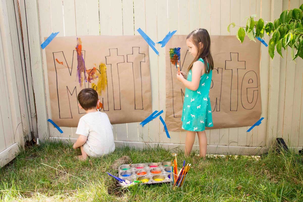 Two kids (2 and 4) standing at large pieces of paper with their names drawn on them beginning to paint.