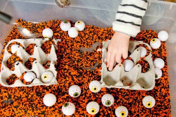 A child's hand reaches into a black and orange rice sensory bin with plastic eye balls.