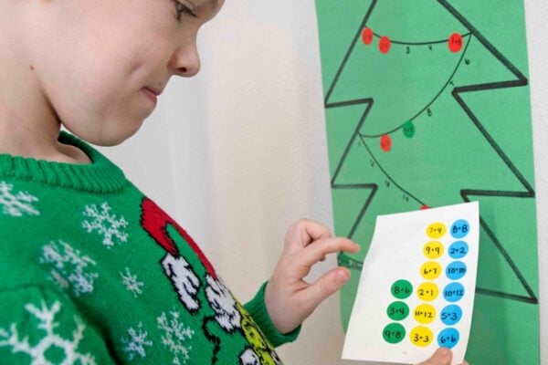 A child studies a sheet of dot stickers with addition facts written on it. A christmas tree with numbers is behind him.