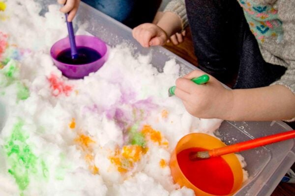 A girl leans over a sensory bin of snow. She's holding a green paint brush and wiping colored water on snow.