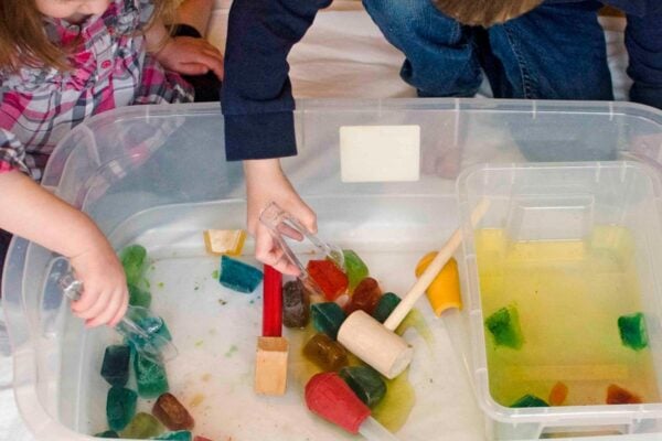 Two children lean over a storage container. There are colored ice cubes in the bin along with tongs, hammers, and basters.