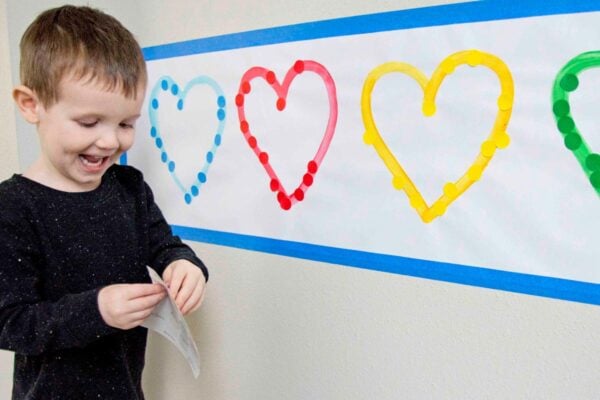 A child smiles while removing a dot sticker from a sheet. On the wall, 4 colored hearts are on white paper. The child is sorting stickers.