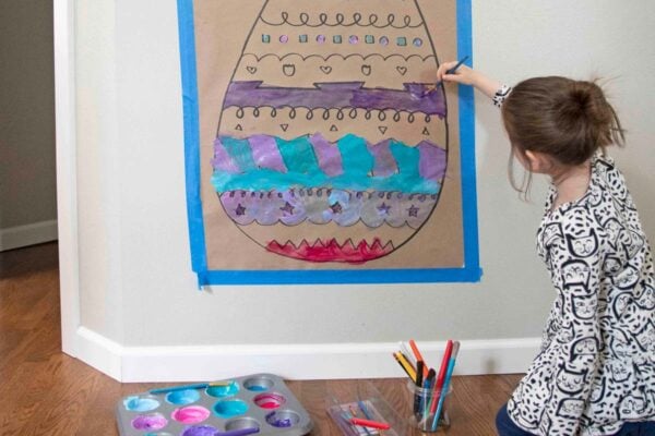 A child reaches up to paint onto a giant Easter egg drawing on Kraft paper. Paint and paint brushes are on the floor in front of her.