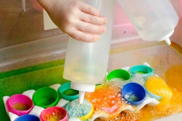 Two children are squeezing condiment bottles full of vinegar. In a tray is half plastic eggs. Colorful bubbles are everywhere.