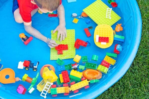A child in a red swim shirt sits in a blue kiddie pool. The pool has water and LEGO Duplo bricks in it.