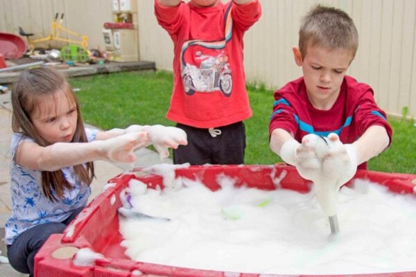 Three kids at a water table outside. The table is full of bubble foam. The kids are covered in it.