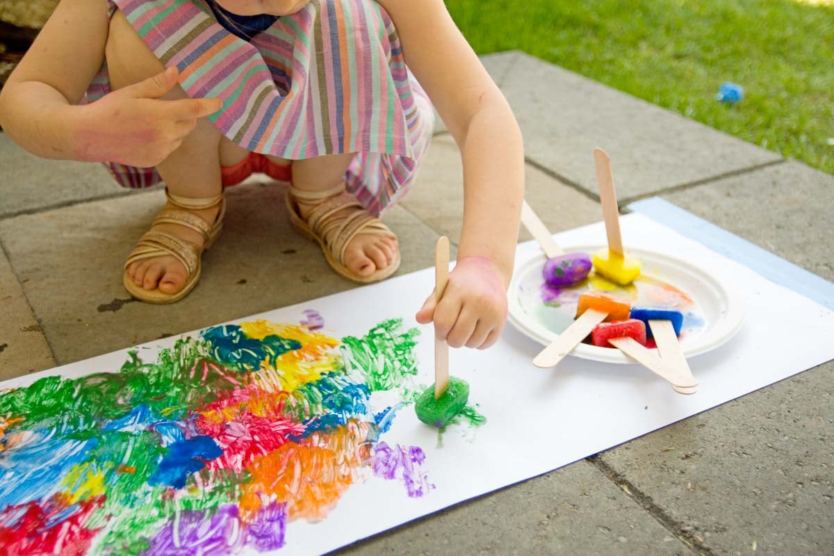 A child holds a craft stick with a green ice cube on it. They are painting with it on white paper. More craft sticks with paint are next to them.