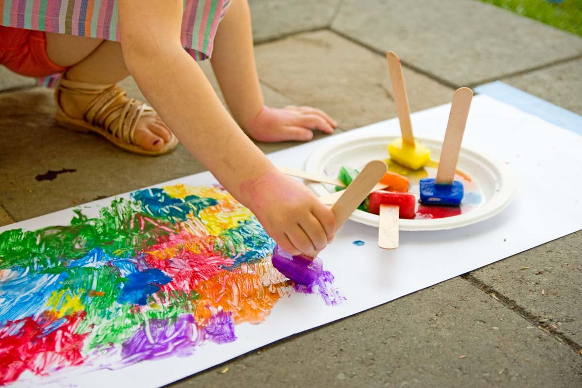 A child holds a craft stick with a blue ice cube on it. They are painting with it on white paper. More craft sticks with paint are next to them.