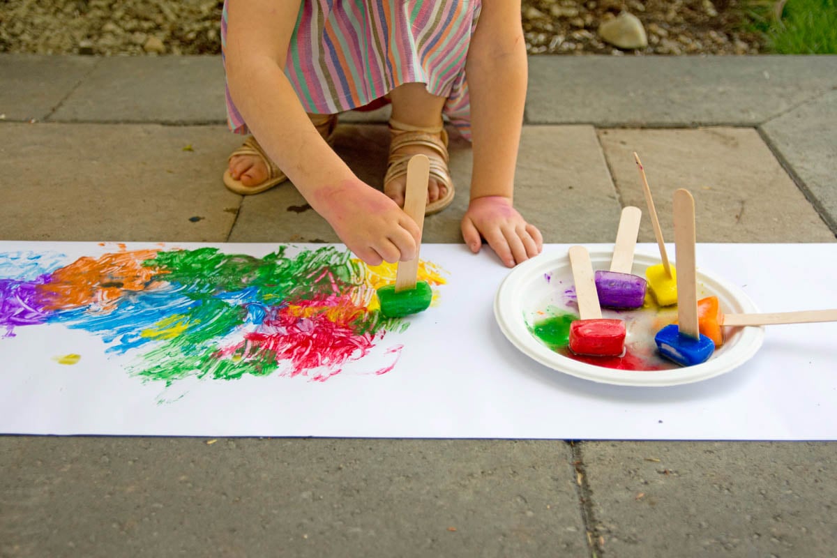 A child holds a craft stick with a blue ice cube on it. They are painting with it on white paper. More craft sticks with paint are next to them.