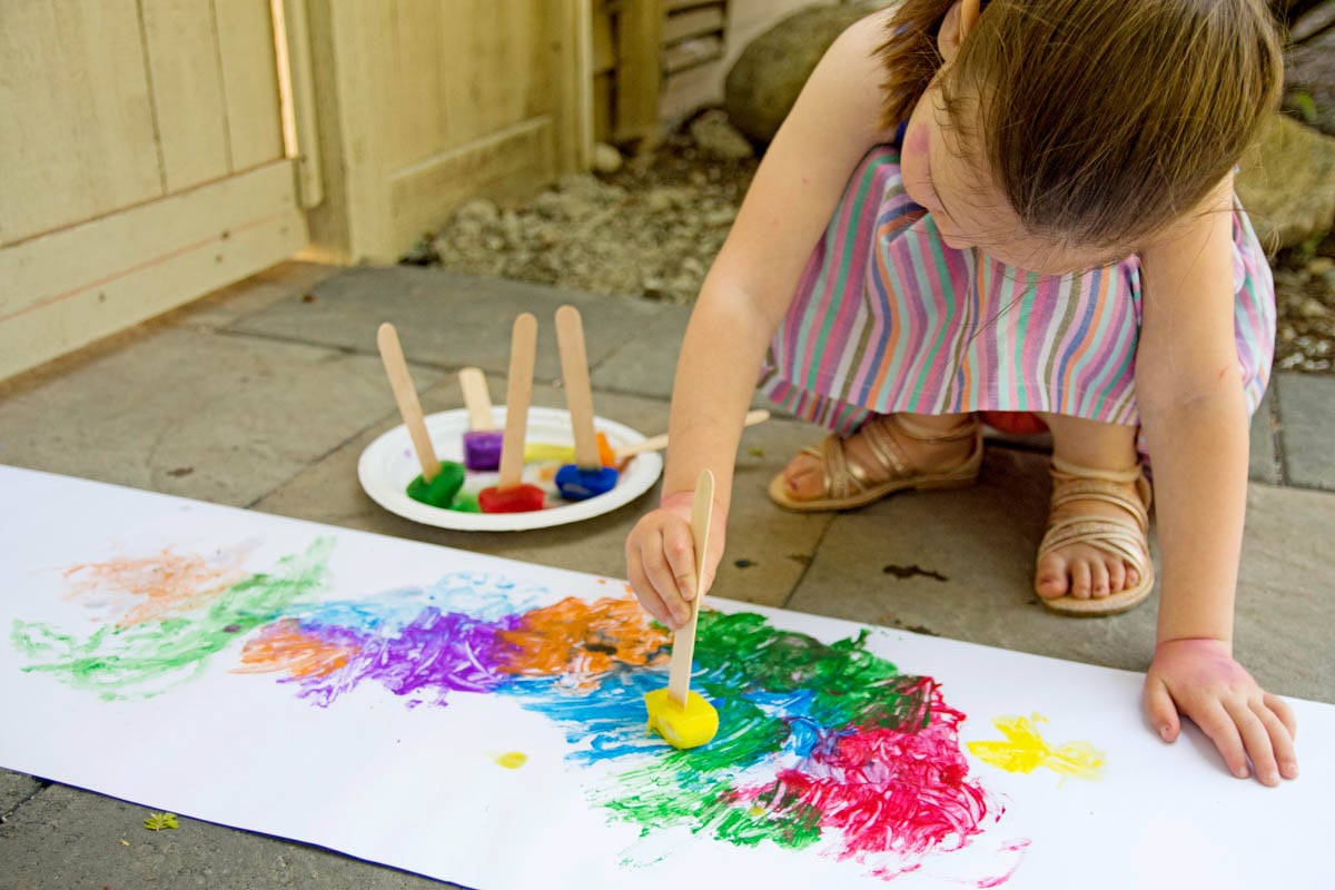 A child holds a craft stick with a yellow ice cube on it. They are painting with it on white paper. More craft sticks with paint are next to them.