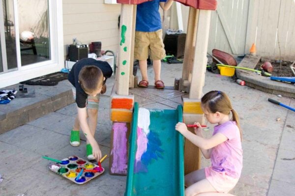Three kids in a backyard are painting their slide and playhouse with washable paint.