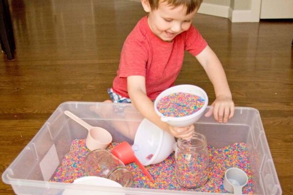 A child smiles while tipping a funnel of rainbow rice into a mason jar. There is a storage container of colorful rice, funnels, and scoops.