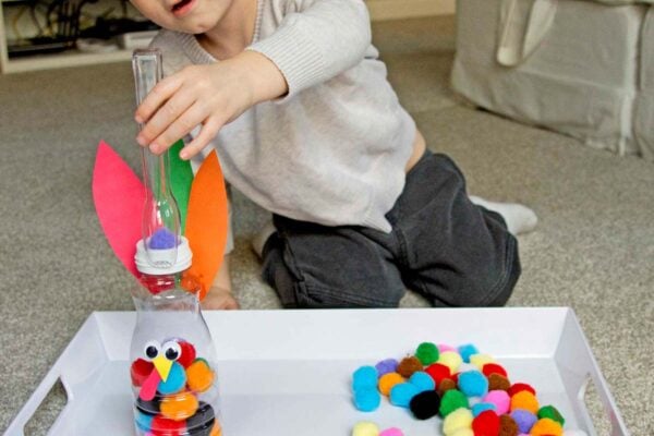 A toddler leans over a creamer bottle that has a turkey face on it. He is placing a pom pom ball into the bottle with tongs.