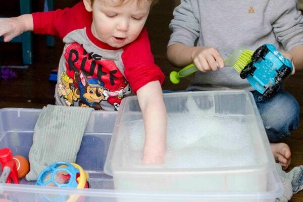 A 1 year old shoves a toy into soapy water as a 4 year old scrubs a toy. They are washing toys together.